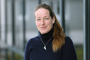 Frau mit langen Haaren und blauen Rollkragenpullover schaut freundlich in die Kamera.