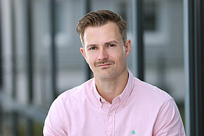 Mann mit kurzen braunen Haaren und Schnauzer ist mit rosafarbenen Hemd gekleidet.