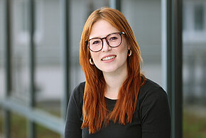 Frau mit langen, rotbraunen Haaren und schwarzem Oberteil, lächelt in die Kamera.