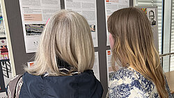 Zwei Frauen betrachten Informationsblätter an einer Pinnwand in einem hellen Raum mit Fensterläden.
