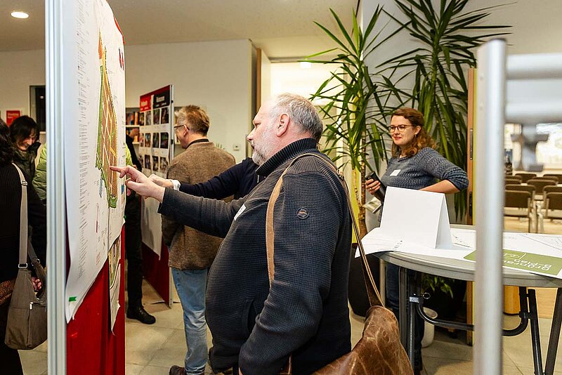 Person steht vor einer Pinnwand und betrachtet Notizen während einer Veranstaltung in einem hellen Raum mit weiteren Teilnehmern im Hintergrund