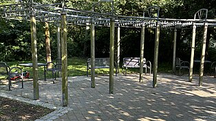 Spielplatz mit Klettergerüst aus Holzpfosten und Sandboden, umgeben von Bäumen.