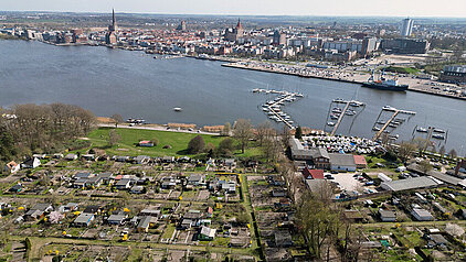 Luftaufnahme eines Gartengeländes mit kleinen Parzellen und Lauben vor der Warnow, im Hintergrund Stadt Rostock mit Kirchtürmen und modernen Gebäuden