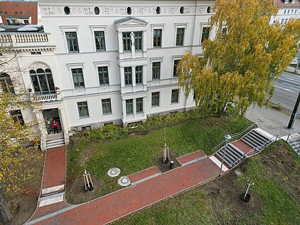 Luftaufnahme eines weißen mehrstöckigen Gebäudes mit rotem Gehweg, Grünfläche und herbstlich gefärbtem Baum