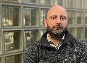 Mann mit Bart in einem grauen Pullover, steht vor einer Wand aus Glasbausteinen, mit neutralem Gesichtsausdruck.