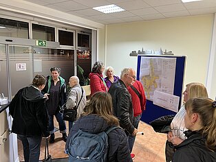 Gruppe von Menschen in einem Raum die diskutieren und einige schauen auf eine Stellwand an der ein Bauplan hängt.