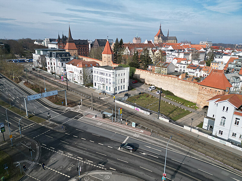 Luftaufnahme von Rostock mit historischen Gebäuden, darunter eine weiße Villa und eine Stadtmauer, umgeben von Straßen und Verkehr.