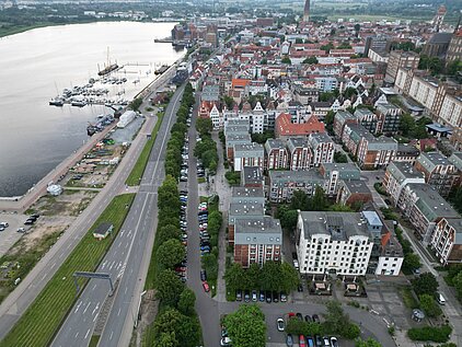 Luftaufnahme einer Stadt mit Geäwsser, mehrspuriger Straße und dicht bebauten Gebäuden.