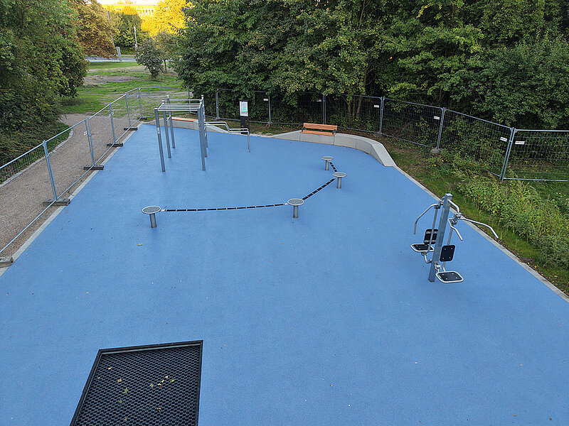 Freiluft-Fitnessbereich mit blauer Bodenfläche, mehreren Trainingsgeräten und einer Bank, umgeben von Bäumen und einem Zaun
