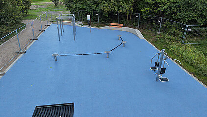 Freiluft-Fitnessbereich mit blauer Bodenfläche, mehreren Trainingsgeräten und einer Bank, umgeben von Bäumen und einem Zaun