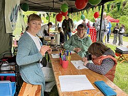 Drei Personen sitzen an einem Holztisch unter einem Zelt, umgeben von bunten Luftballons und Wimpelketten, eine Person malt, zwei andere lächeln in die Kamera, im Hintergrund weitere Menschen auf einer grünen Wiese