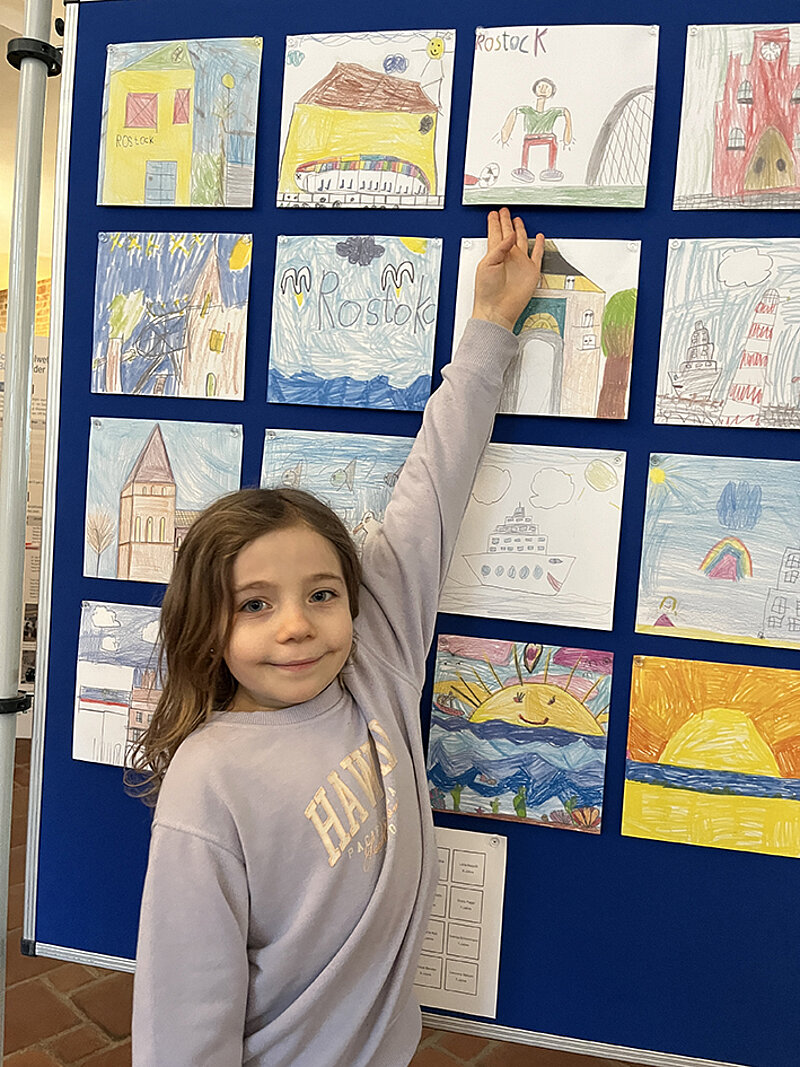 Kind zeigt auf eine Wand mit mehreren bunten Kinderzeichnungen und ihrem Bild mit einem Fußballspieler