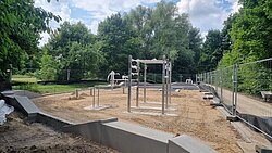 Freiluft-Fitnessbereich mit Metall-Klimmzugstangen und Sandboden, umgeben von Bäumen und Bauzaun