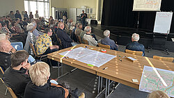 Gruppe von Menschen sitzt in einem Raum und hört einem Vortrag zu, auf einer Leinwand wird ein Plan oder eine Karte gezeigt, Tische mit weiteren Karten und Materialien im Vordergrund
