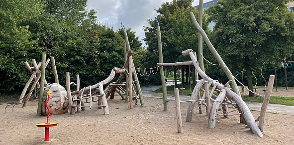 Spielplatz mit Klettergerüst aus naturbelassenem Holz in Form von Dinosauriern und Sandboden, im Vordergrund ein kleines rotes Karussell