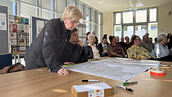 Personen in einem hellen Raum, eine ältere Frau beugt sich über einen Tisch mit großen Plänen, im Hintergrund sitzen weitere Menschen und hören zu