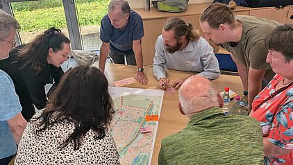 Gruppe von Menschen, die um einen Tisch versammelt sind und gemeinsam an einem Plan oder einer Karte arbeiten. Einige Personen zeigen auf den Plan, während andere aufmerksam zuhören.