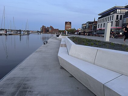 Uferpromenade mit modernen Sitzbänken aus Beton, Wasser links und mehrstöckigen Gebäuden rechts
