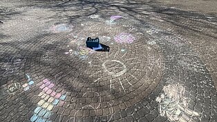 Bunte Kreidezeichnungen auf gepflastertem Boden, darunter Kreise, Muster und ein kleiner blauer Kasten in der Mitte