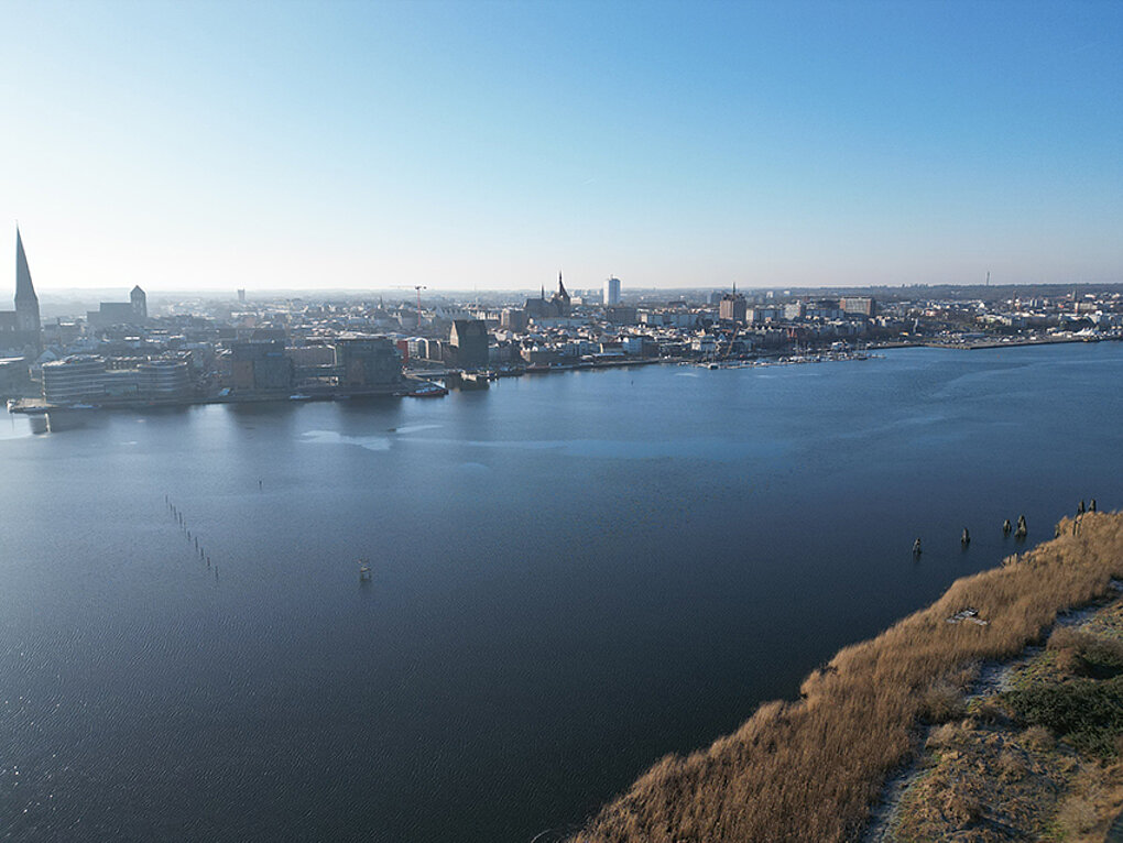 Stadtpanorama an der Unterwarnow im Vordergrund und Gebäuden am Ufer unter klarem Himmel
