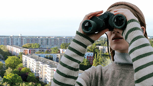 Junge Frau mit gestreiftem Pullover, die durch ein Fernglas blickt. Im Hintergrund sind Wohngebäude und Bäume sichtbar.