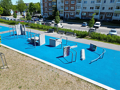 Blauer Outdoor-Fitnessbereich mit verschiedenen Trainingsgeräten auf Asphalt, umgeben von Wohngebäuden und Grünflächen