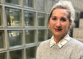 Frau mit blonden, schulterlangen Haaren und einem freundlichen Lächeln, trägt einen grauen Pullover mit einem weißen Kragen. Im Hintergrund sind Glasbausteine sichtbar.