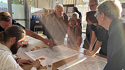 Acht Personen diskutieren und zeigen auf große Stadtpläne auf einem Tisch in einem hellen Raum mit großen Fenstern