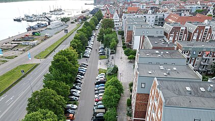 Blick von oben auf eine Straße mit parkenden Autos und Bäumen entlang eines Gewässers in einer Stadt.