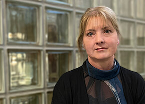 Porträt einer Frau mit blonden Haaren, die in einem schwarzen Oberteil und einem blauen Schal gekleidet ist, vor einer Glaswand.