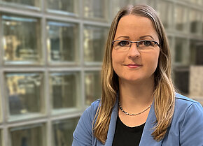 Frau mit langen Haaren und Brille, trägt einen blauen Blazer. Sie steht vor einer modernen Glaswand.