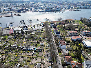 Luftaufnahme mit Gärten und Häusern an einem Flussufer der Warnow mit Bootsanlegern und Stadt Rostock im Hintergrund
