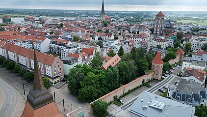 Luftaufnahme der Rostocker Innenstadt mit roten Dächern, zwei markanten Kirchtürmen und einem Fluss im Hintergrund unter bewölktem Himmel