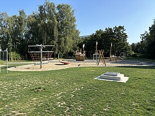 Spielplatz mit verschiedenen Spielgeräten, umgeben von Bäumen und Rasenfläche, bei sonnigem Wetter.