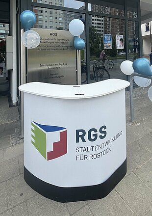 Empfangstresen vor einem Gebäude mit dem Logo der Firma RDS, dekoriert mit blauen und grauen Luftballons.