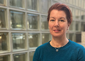 Frau mit kurzen, roten Haaren in blauer Strickjacke vor einer Wand aus Glasbausteinen, lächelt freundlich.