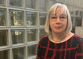 Frau mit schulterlangen Haaren und Brille, trägt ein rot-schwarzes, geometrisches Oberteil. Sie steht vor einer Wand aus Glasbausteinen.