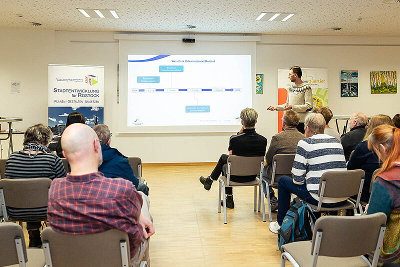 Mehrere Personen sitzen in einem Raum und schauen auf eine Präsentation auf einer großen Leinwand