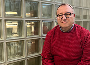 Mann mit Brille und rotem Pullover sitzt vor einer Wand aus Glasbausteinen. Er hat einen neutralen Gesichtsausdruck.