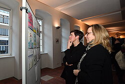 Zwei Personen betrachten eine Wand mit mehreren Aushängen in einem Innenraum mit Säulen