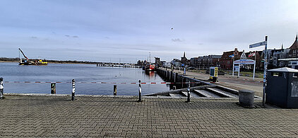 Blick auf den Stadthafen mit ruhigem Wasser, mehreren Booten und Gebäuden am Ufer unter bewölktem Himmel