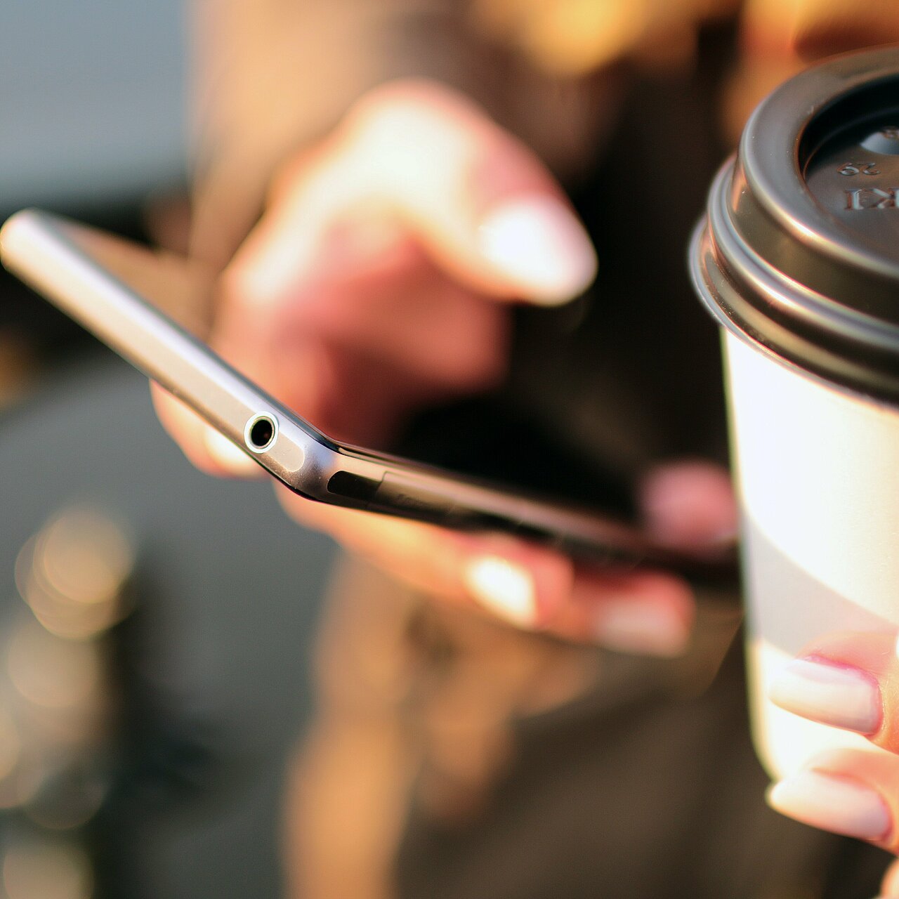 Eine Hand hält ein Smartphone und eine Kaffeetasse. Der Hintergrund zeigt unscharfe Elemente, die auf eine entspannte Umgebung hinweisen.