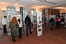 Mehrere Personen betrachten in einem Ausstellungsraum auf Ständern präsentierte großformatige Fotografien oder Plakate