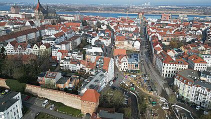 Luftaufnahme von Rostock mit roten Dächern, historischen Gebäuden und der Unterwarnow im Hintergrund.