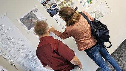 Zwei Personen betrachten Pläne an einer Wand in einem Ausstellungsraum. Eine Person trägt eine braune Jacke und hat eine schwarze Tasche, die andere Person trägt ein rotes Hemd.
