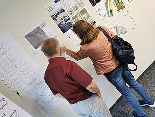 Zwei Personen betrachten Pläne an einer Wand in einem Ausstellungsraum. Eine Person trägt eine braune Jacke und hat eine schwarze Tasche, die andere Person trägt ein rotes Hemd.