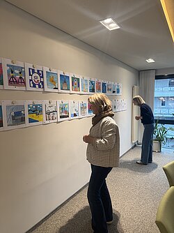 Zwei Personen betrachten bunte Bilder, die in einer Reihe an einer weißen Wand hängen.