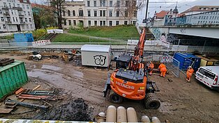 Bauarbeiter in orangefarbener Kleidung arbeiten auf einer Baustelle mit schwerem Gerät und Baucontainern. 