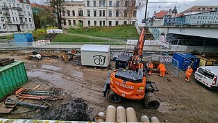 Bauarbeiter in orangefarbener Kleidung arbeiten auf einer Baustelle mit schwerem Gerät und Baucontainern. 
