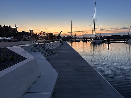Uferpromenade mit Sitzstufen, Segelbooten im Wasser und Sonnenuntergang am Horizont